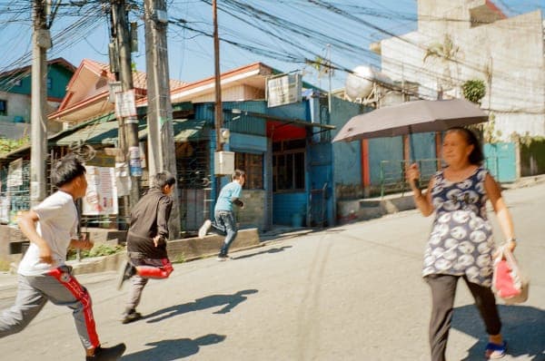 A photograph of three children running in a staggered formation, their speed conveyed by slight motion blur. In the foreground, a woman walks with her umbrella open, shielding herself from the blazing sun.